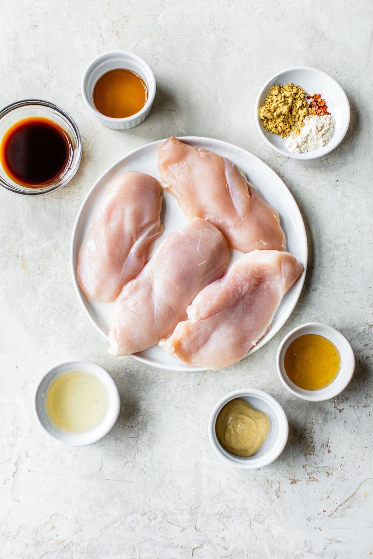 Raw chicken breast on a plate with ingredients for marinade in small bowls.