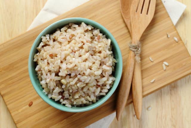 brown rice sides in a sea foam green bowl on top of a cutting board with wooden utensils next to it