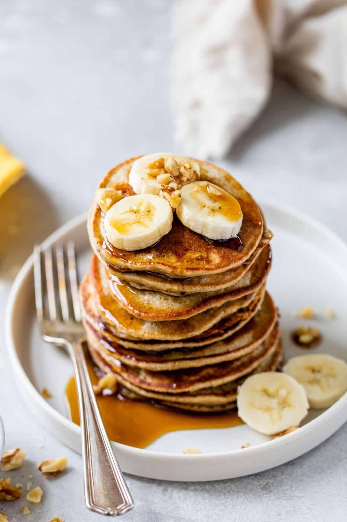 a stack of banana oatmeal pancakes topped with banana slices, chopped nuts and maple syrup 