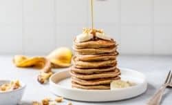 pouring maple syrup over a stack of pancakes on a white plate