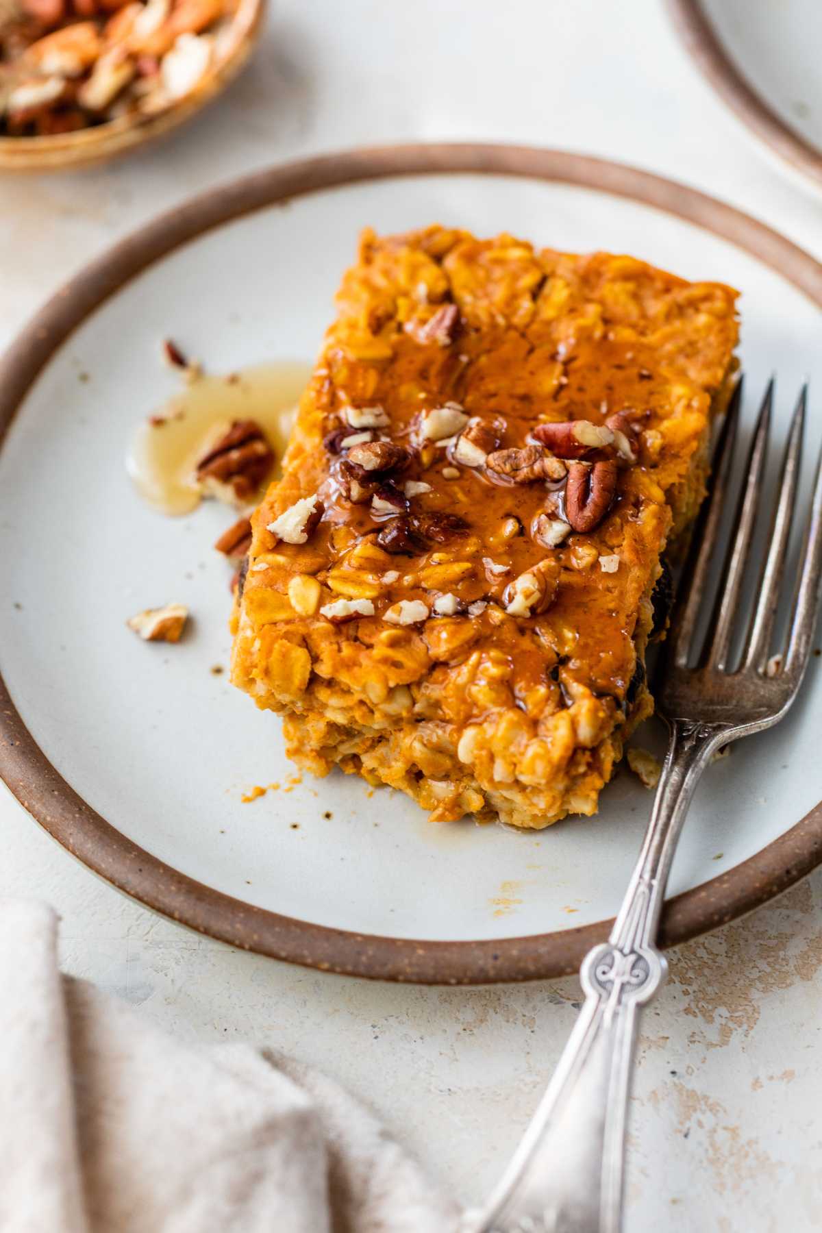Slice of baked pumpkin oatmeal topped with syrup and chopped pecans on a white plate.