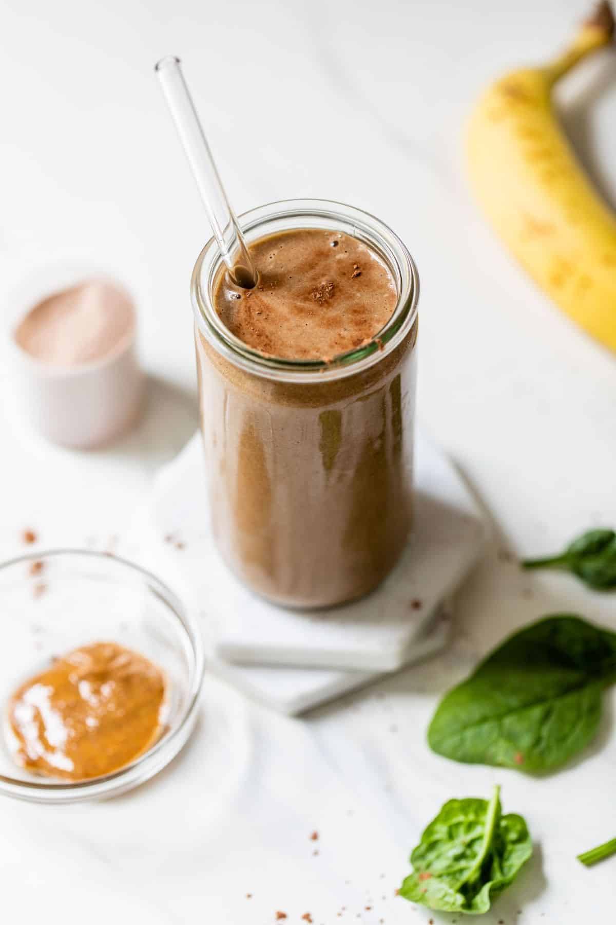 chocolate protein shake in a glass near a banana and spinach leaves