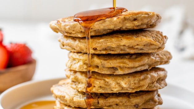 oatmeal pancakes stacked on white plate and drizzled with maple syrup