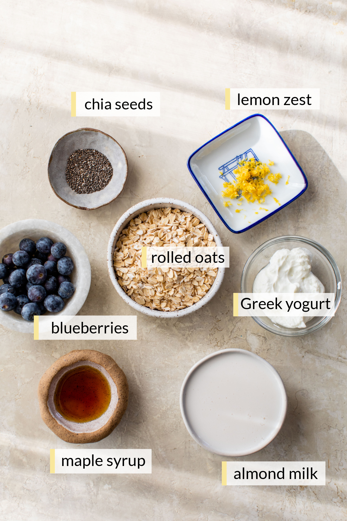 Blueberries, lemon zest, oats, Greek yogurt, maple syrup, chia seeds and almond milk divided into small bowls.