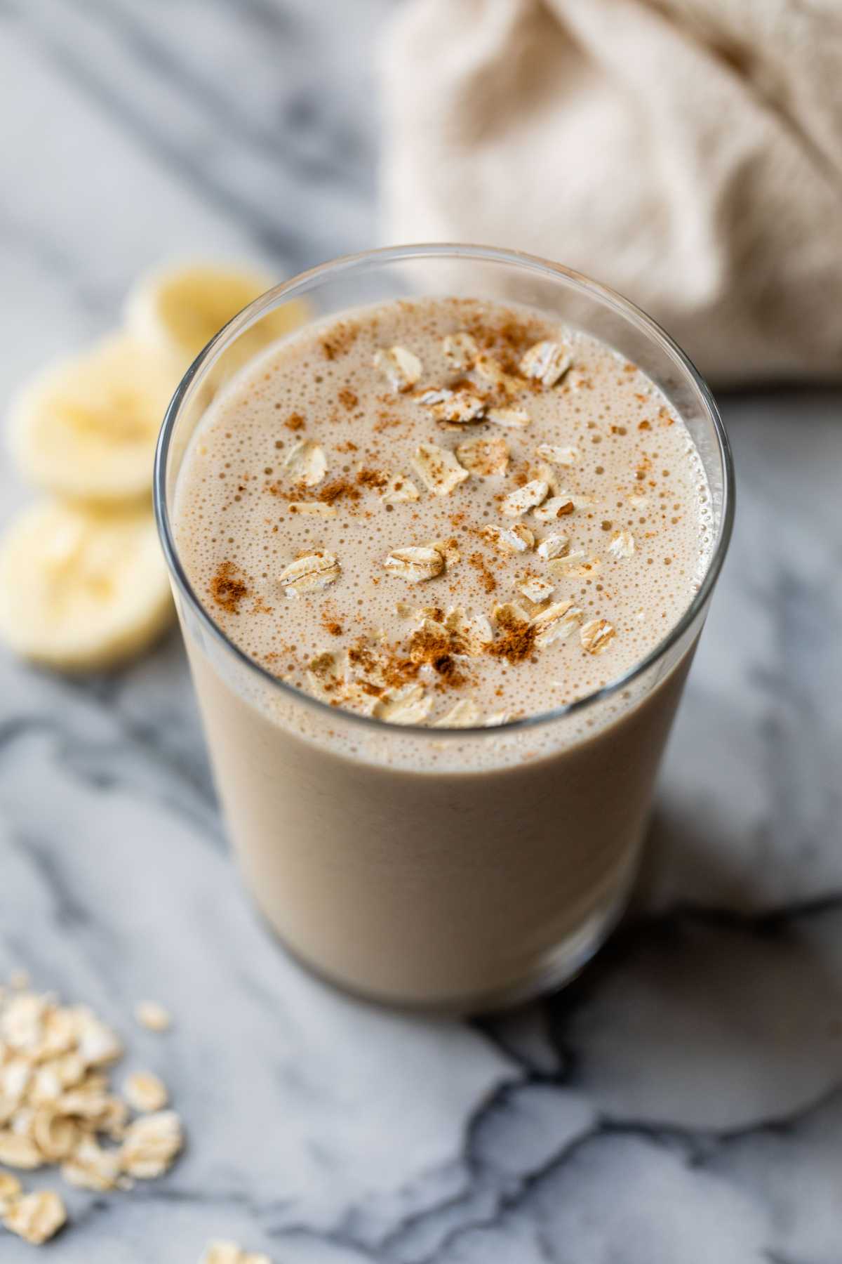 Breakfast smoothie in a glass near banana slices and rolled oats.