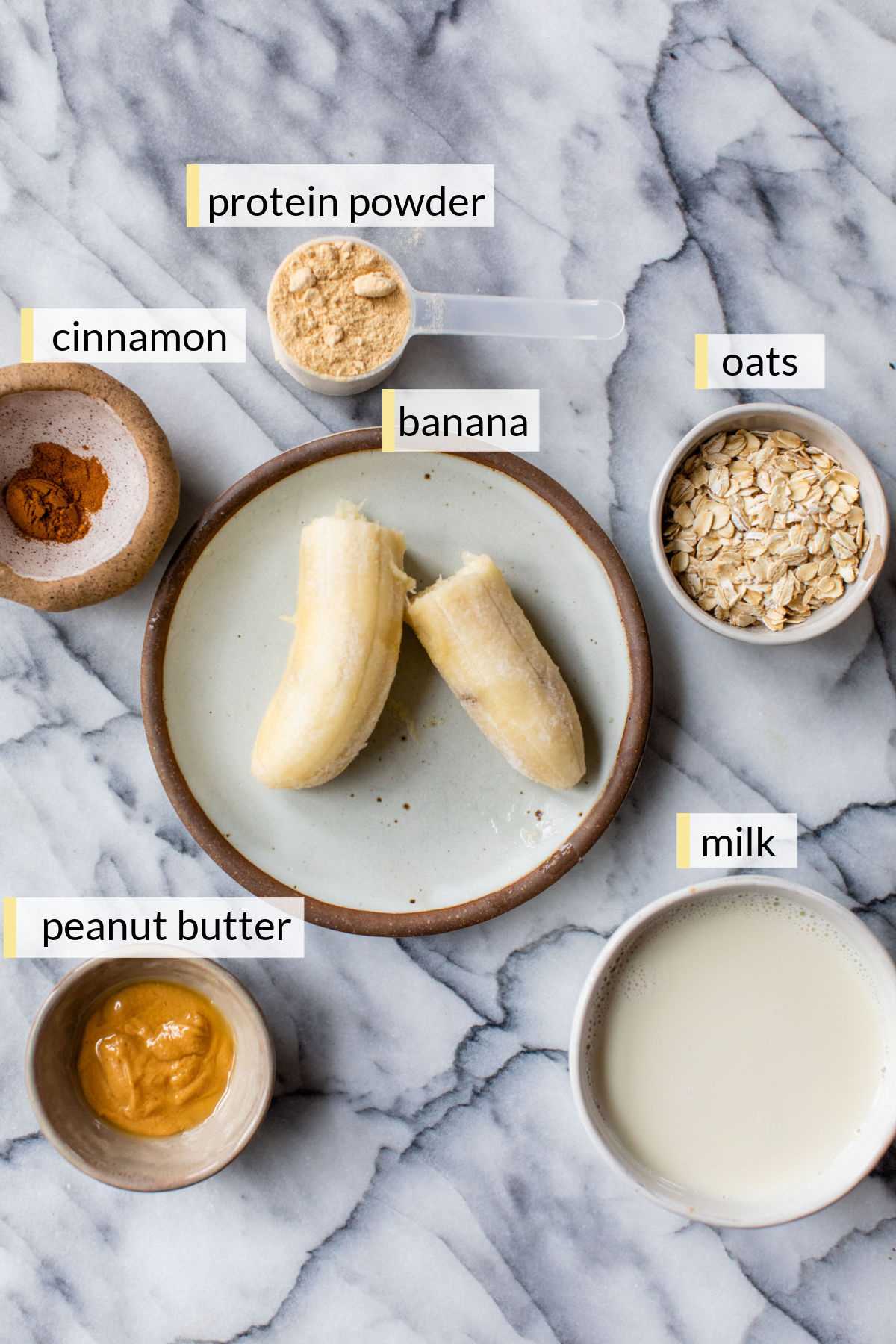 Banana on a plate near oats, milk, peanut butter and protein powder in small bowls.