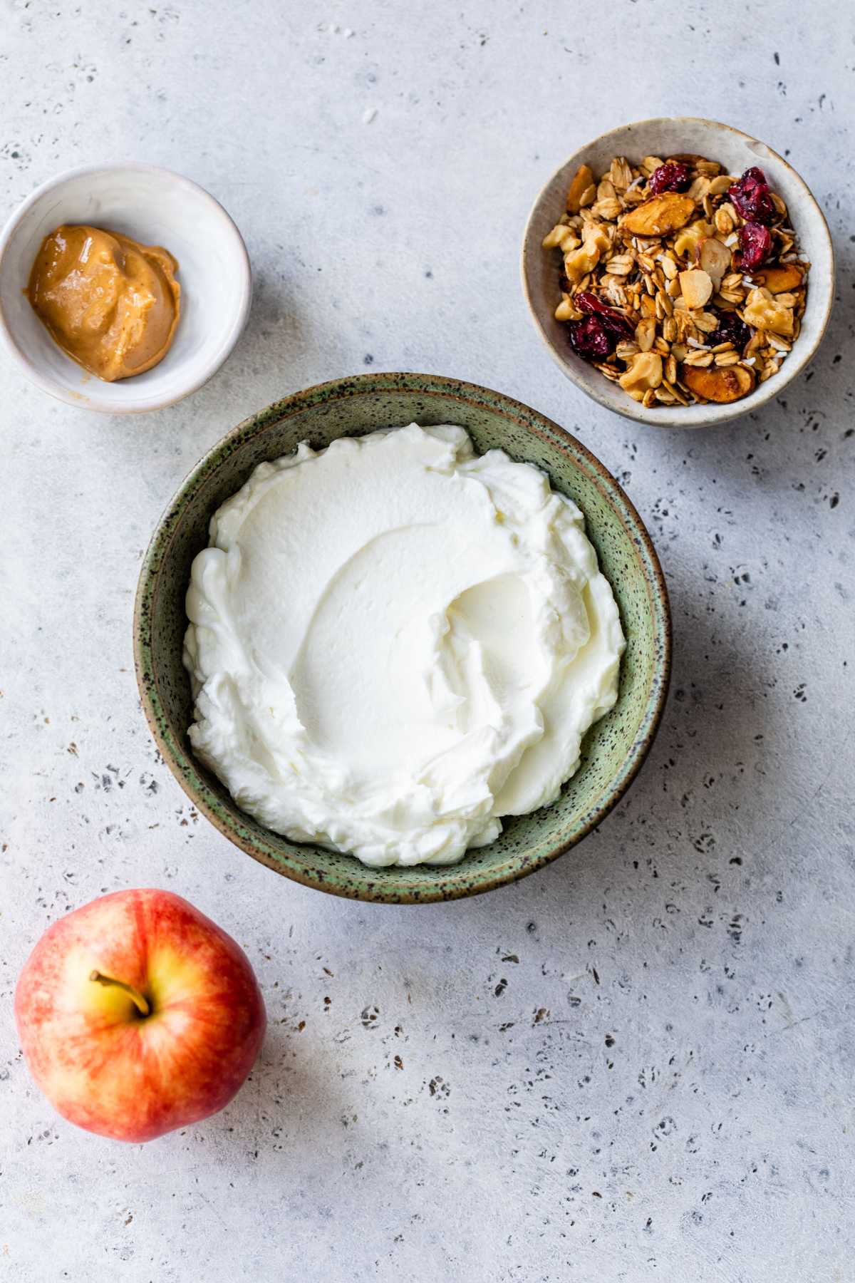 Greek yogurt in a bowl near an apple, a small bowl of peanut butter and a bowl of granola.