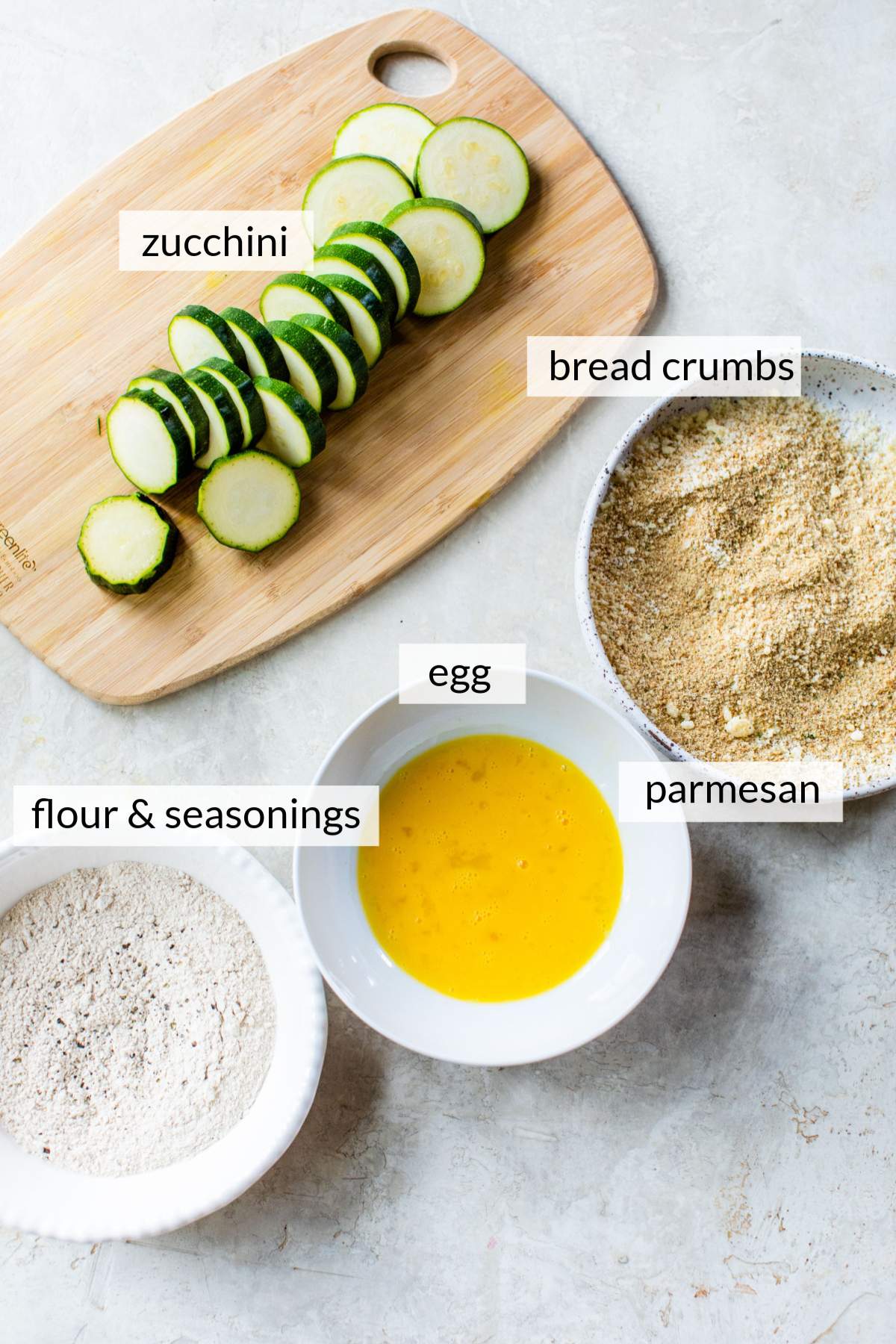 Sliced zucchini on a cutting board near bowls of breadcrumbs, whisked egg and flour.