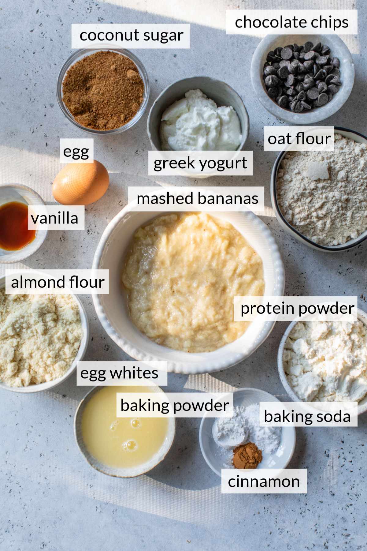 Mashed bananas in a bowl near ingredients like chocolate chips, egg whites, flour and vanilla.